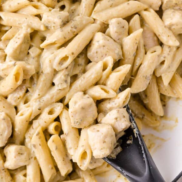 Close-up of creamy penne pasta with pieces of chicken, mixed with a light sauce and herbs, being stirred with a black serving spoon.
