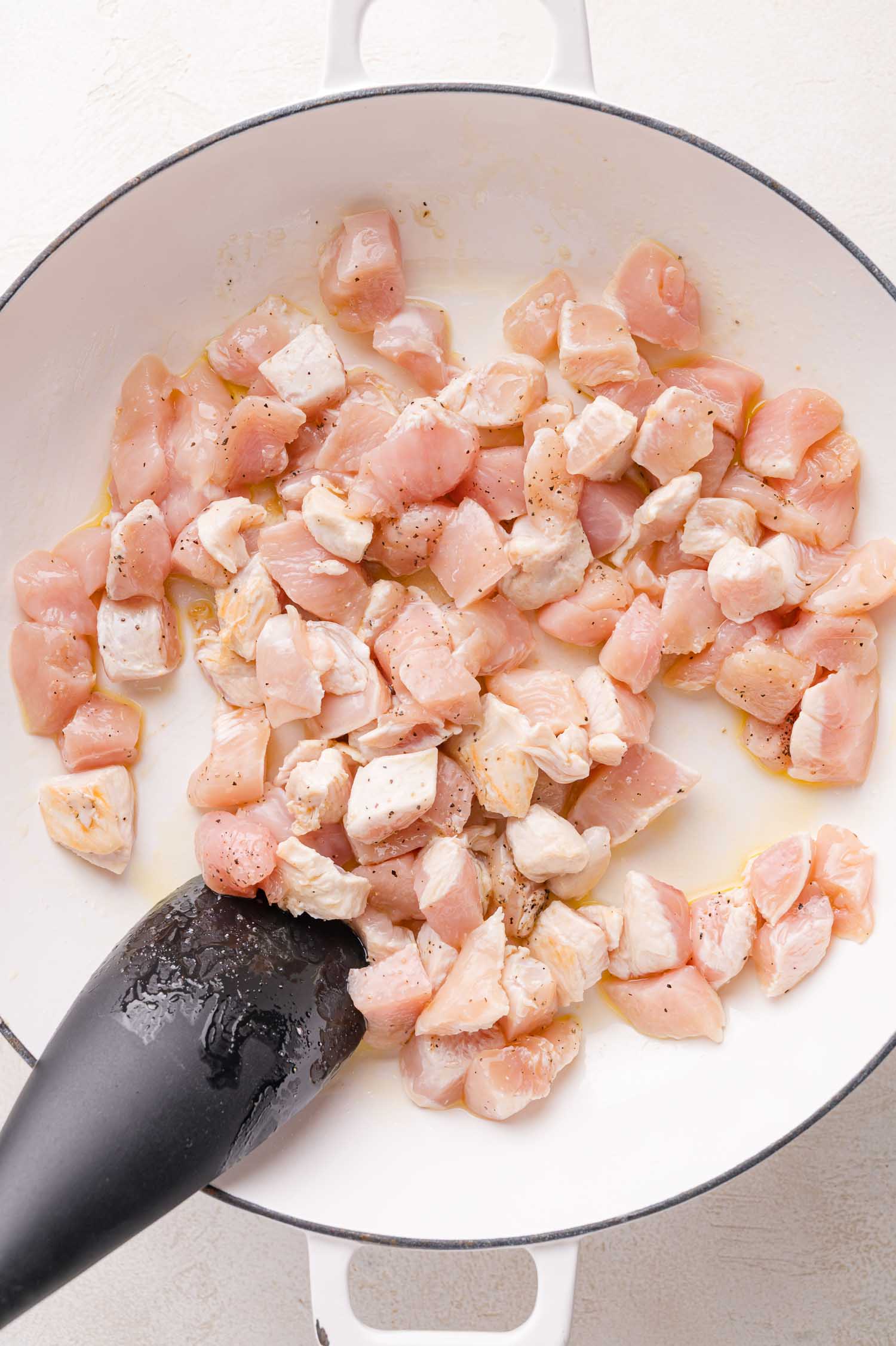 Diced raw chicken pieces being cooked in a white skillet with a black spatula.