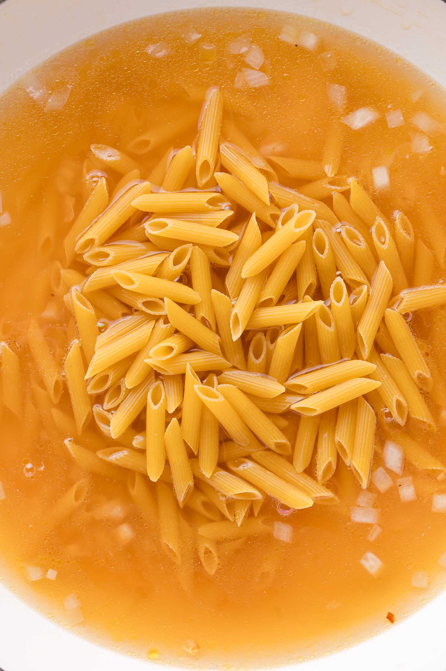 Uncooked penne pasta in a pot with broth and chopped onions.