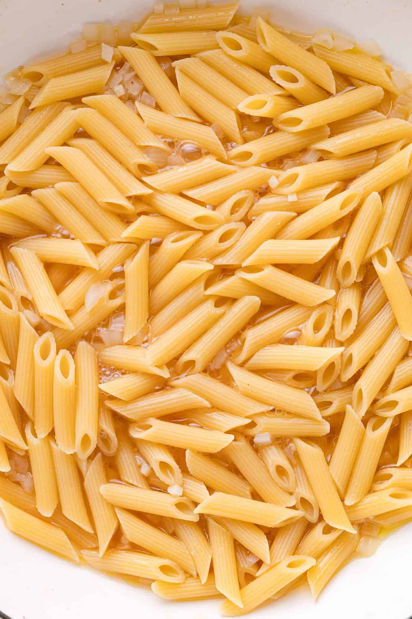 Uncooked penne pasta in a pot with chopped onions and water.