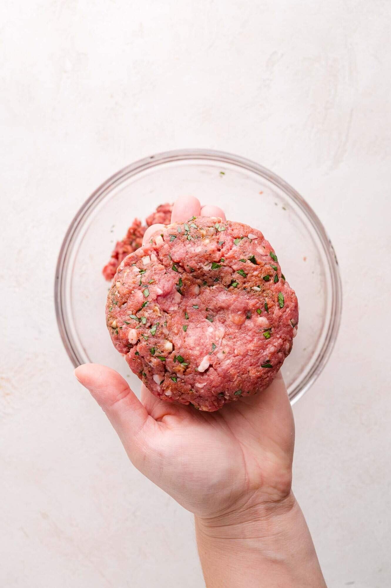 A hand holds a raw, seasoned ground lamb patty over a glass bowl on a light-colored surface.