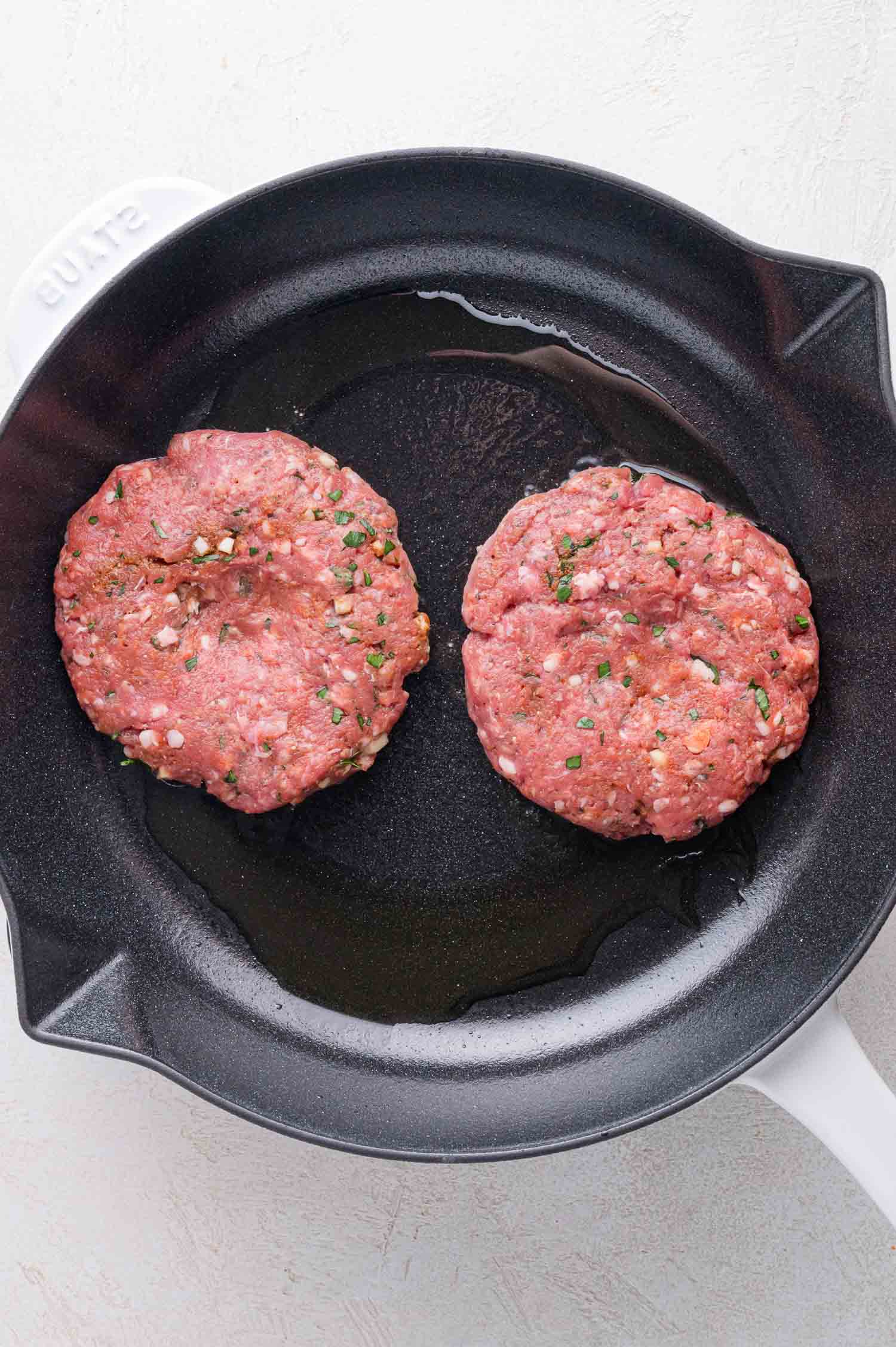 Two raw seasoned burger patties are cooking in a black skillet with a small amount of oil.