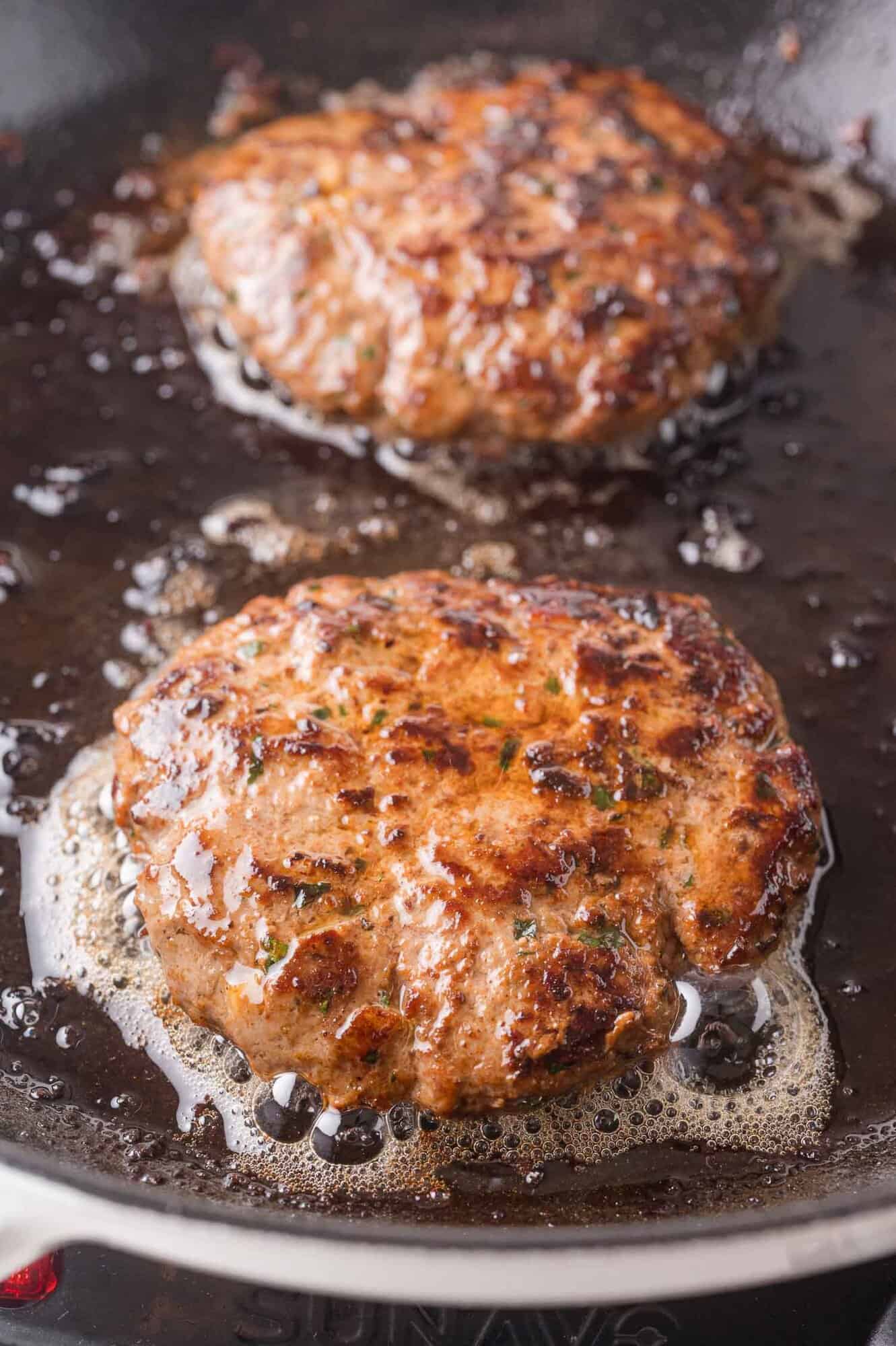 Two browned ground lamb patties sizzling in a frying pan with bubbling oil.