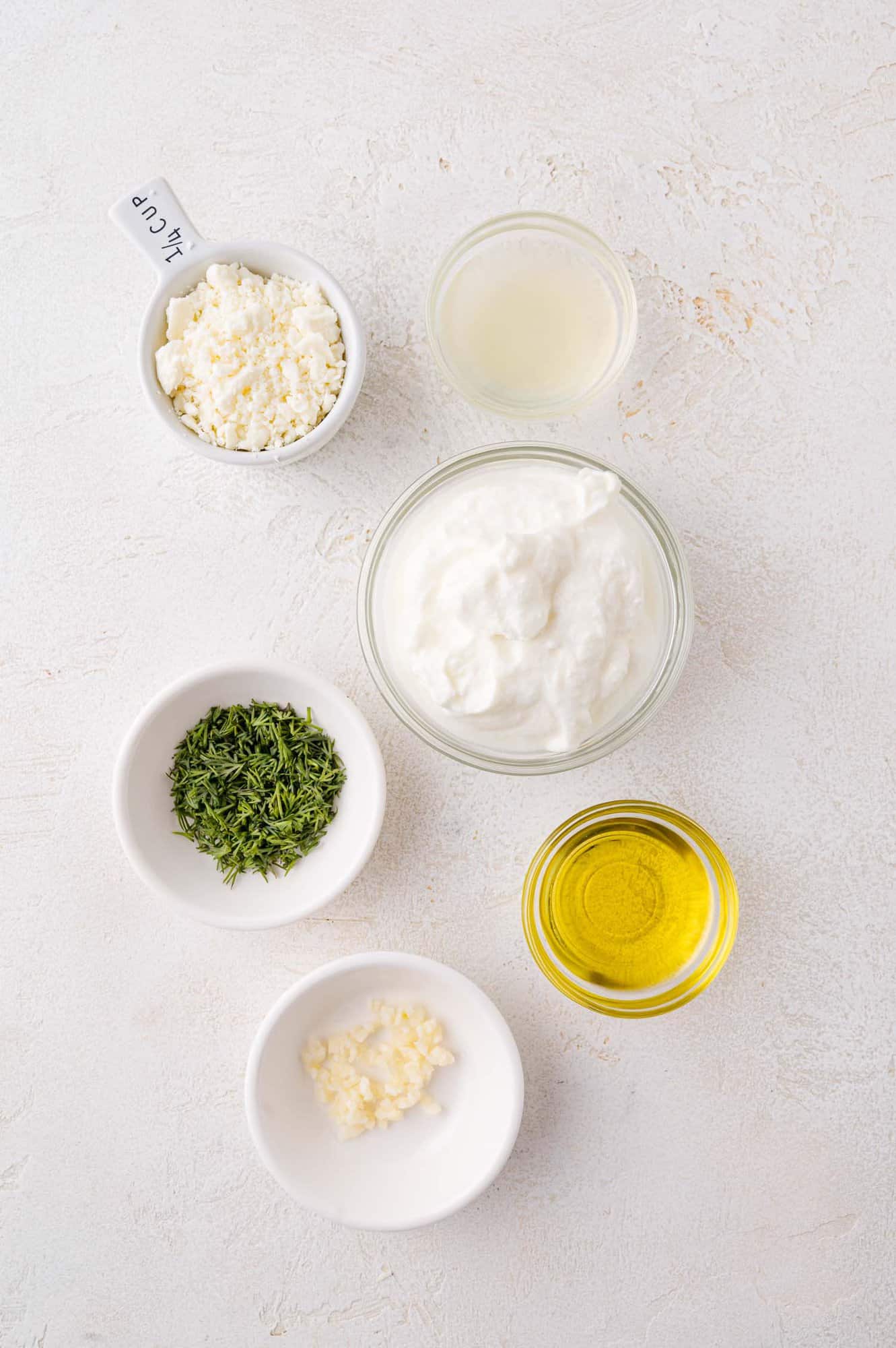 Five small bowls containing feta cheese, lemon juice, Greek yogurt, chopped herbs, olive oil, and minced garlic are arranged on a light textured surface.