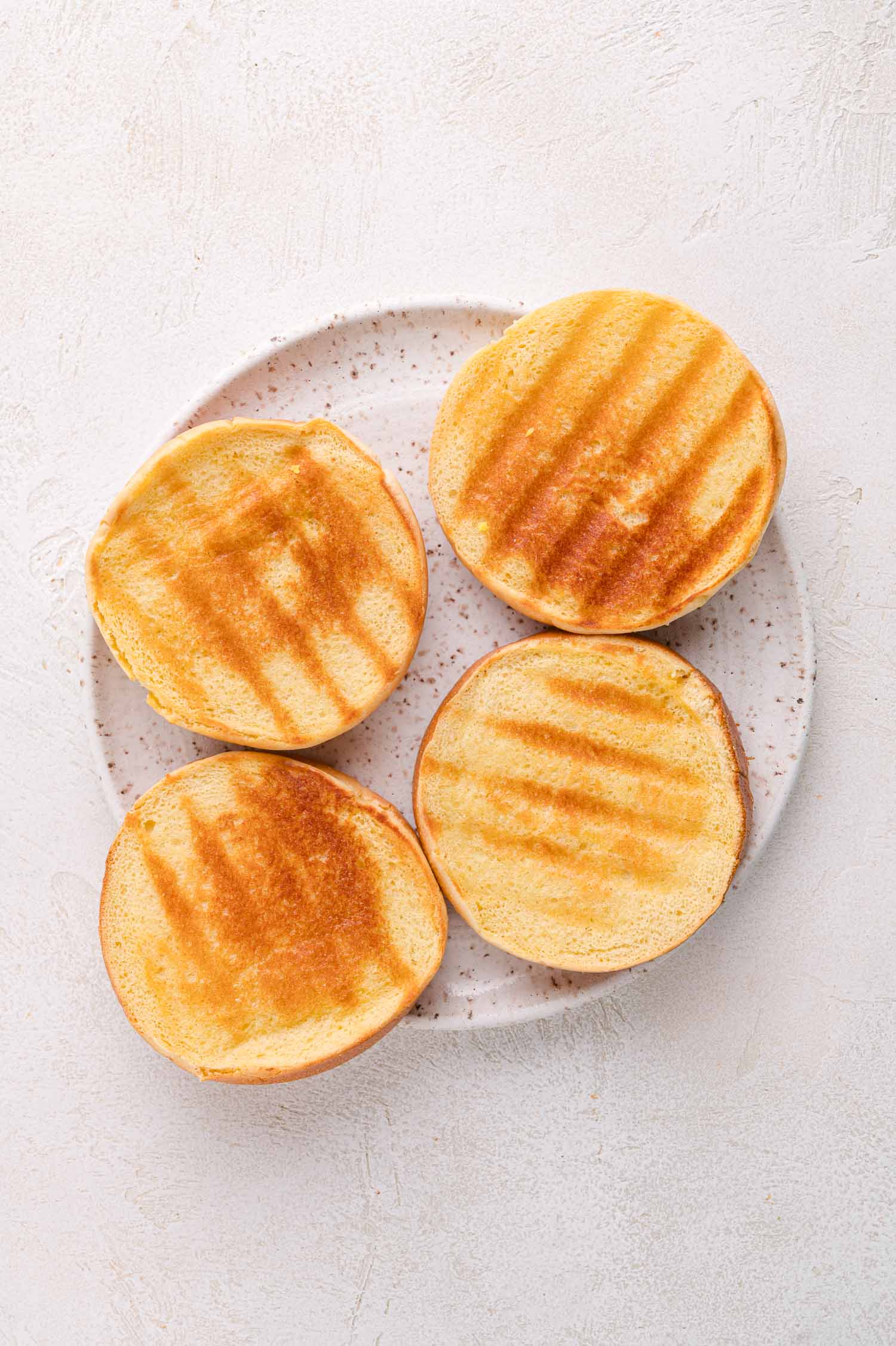 A plate with four toasted burger bun halves, showing grill marks on a light textured surface.