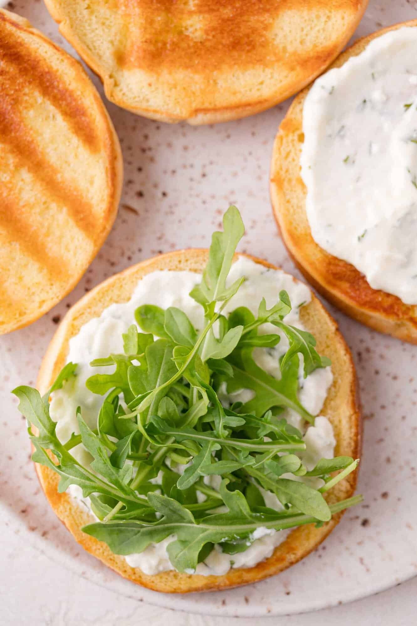 A toasted sandwich bun topped with a white spread and fresh arugula, next to other toasted buns with spread, on a speckled plate.