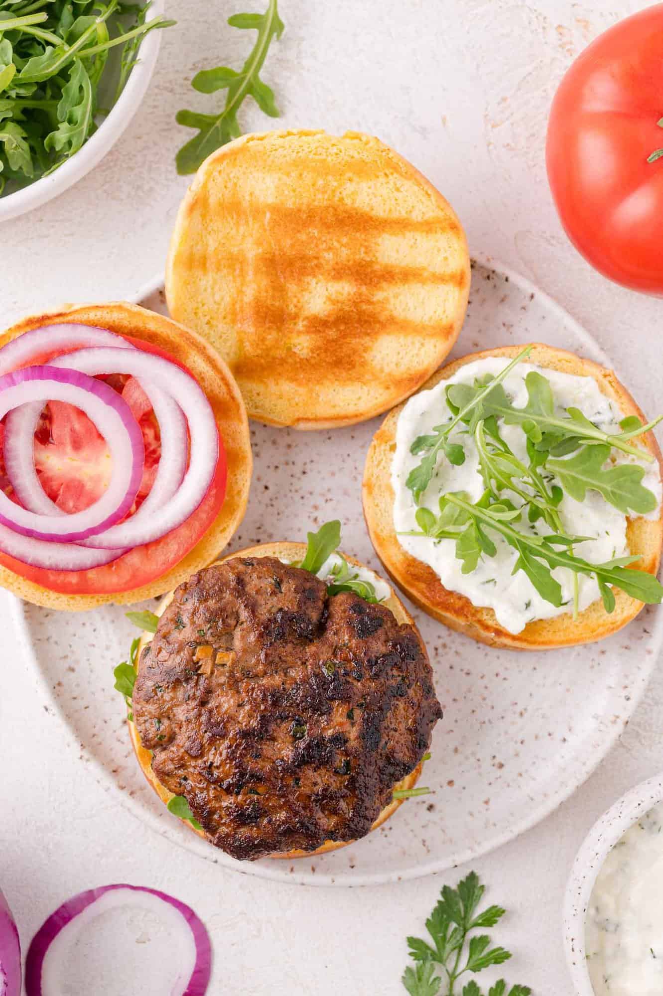 Open-faced burger on a plate with a grilled patty, arugula, sauce, tomato slices, and red onion on a toasted bun. Nearby are arugula, tomato, and onion slices.