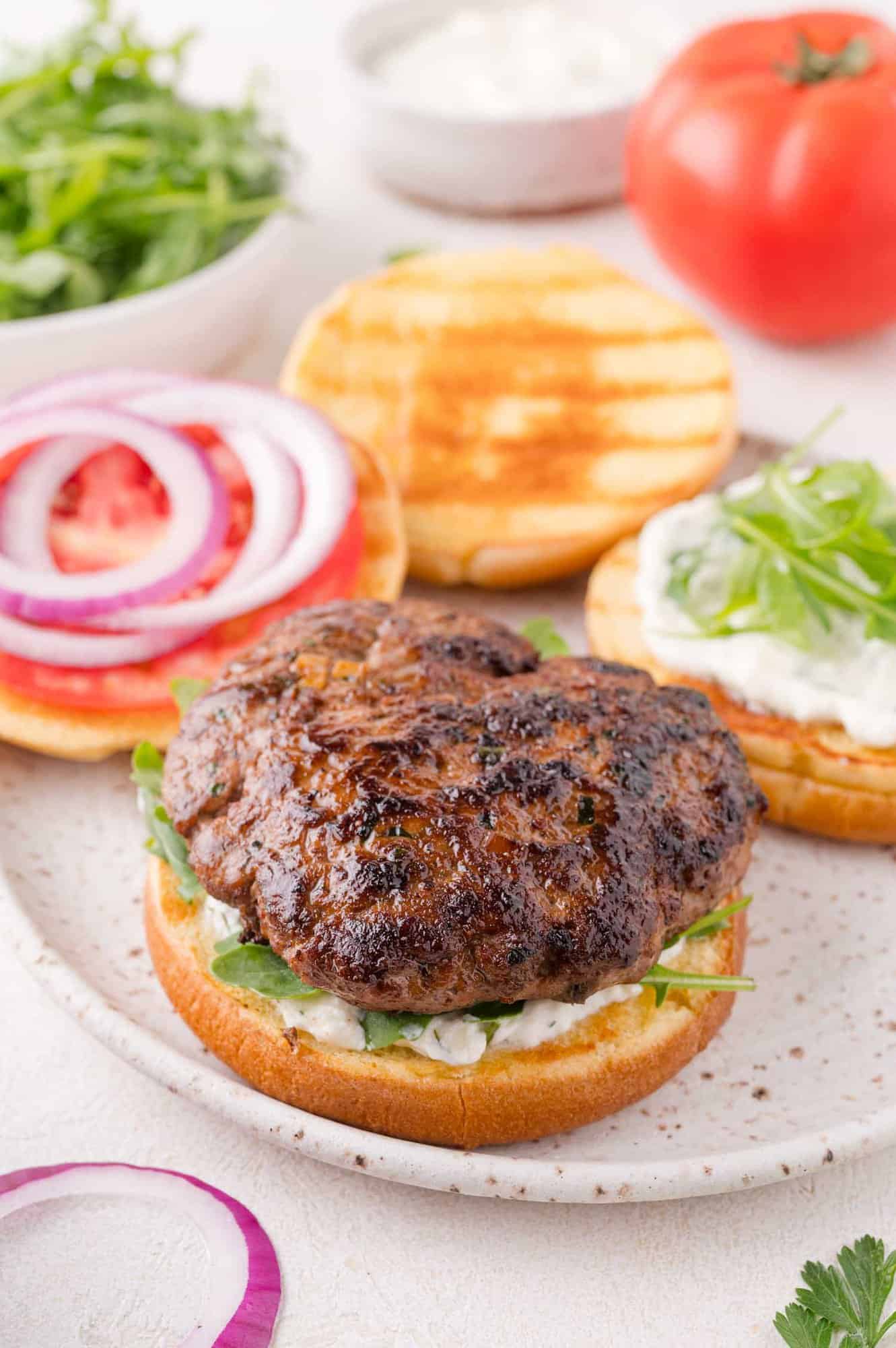 A grilled lamb burger patty sits on a bun with greens and sauce, alongside sliced tomato, red onion, and another bun half with sauce and greens, on a plate with fresh ingredients nearby.