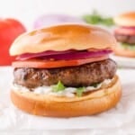 A lamb burger with sliced tomato, red onion, lettuce, and white sauce in a toasted bun, shown on a white surface.