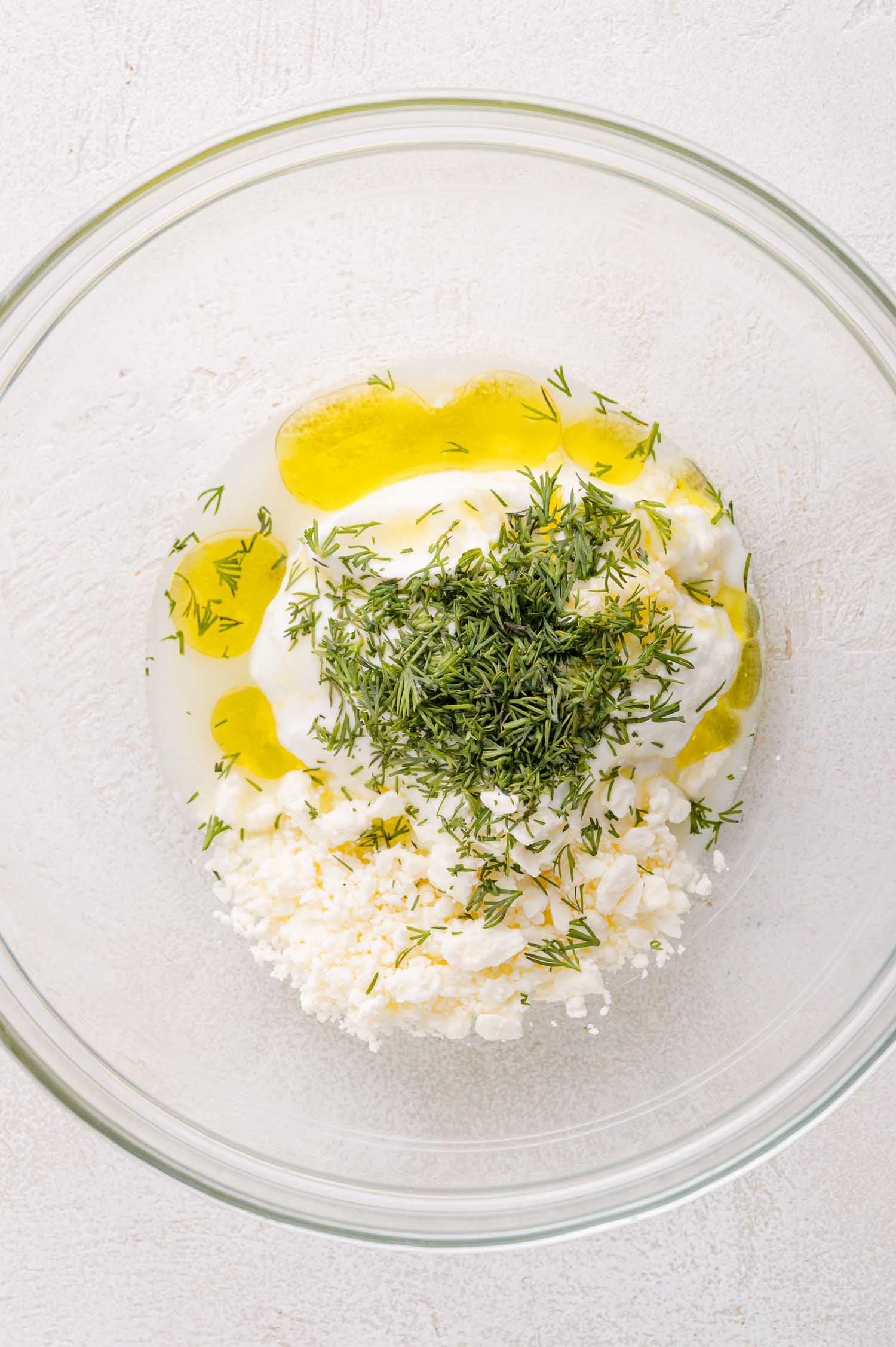 A glass bowl containing feta cheese, plain yogurt, chopped dill, olive oil, and lemon juice on a light surface.