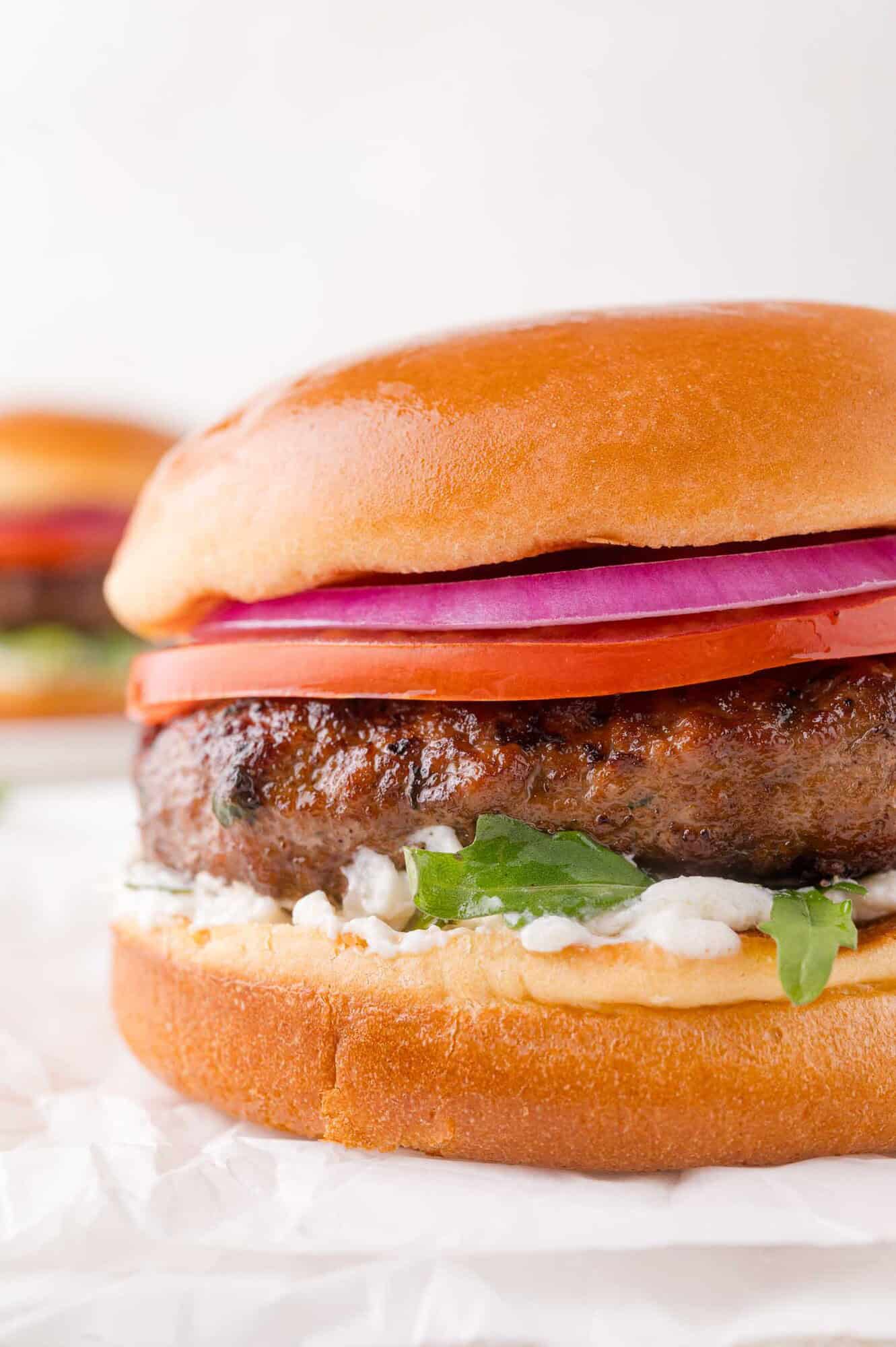 A close-up of a lamb burger with sliced red onion, tomato, lettuce, and sauce in a glossy bun on white parchment paper.