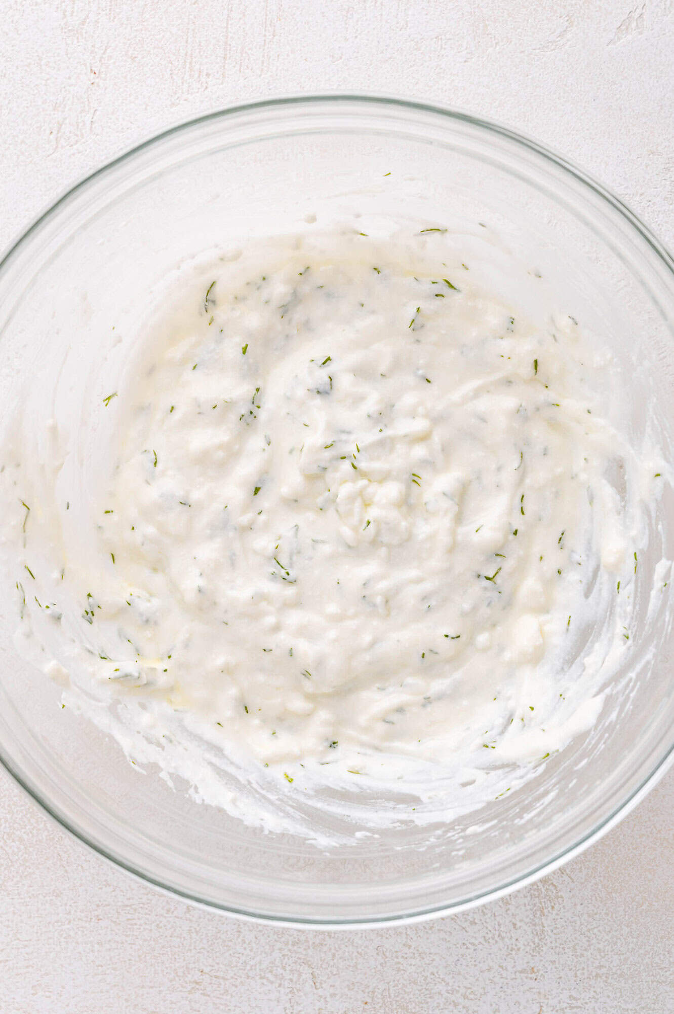 A glass bowl containing a creamy white mixture with visible green herb flecks on a light surface.