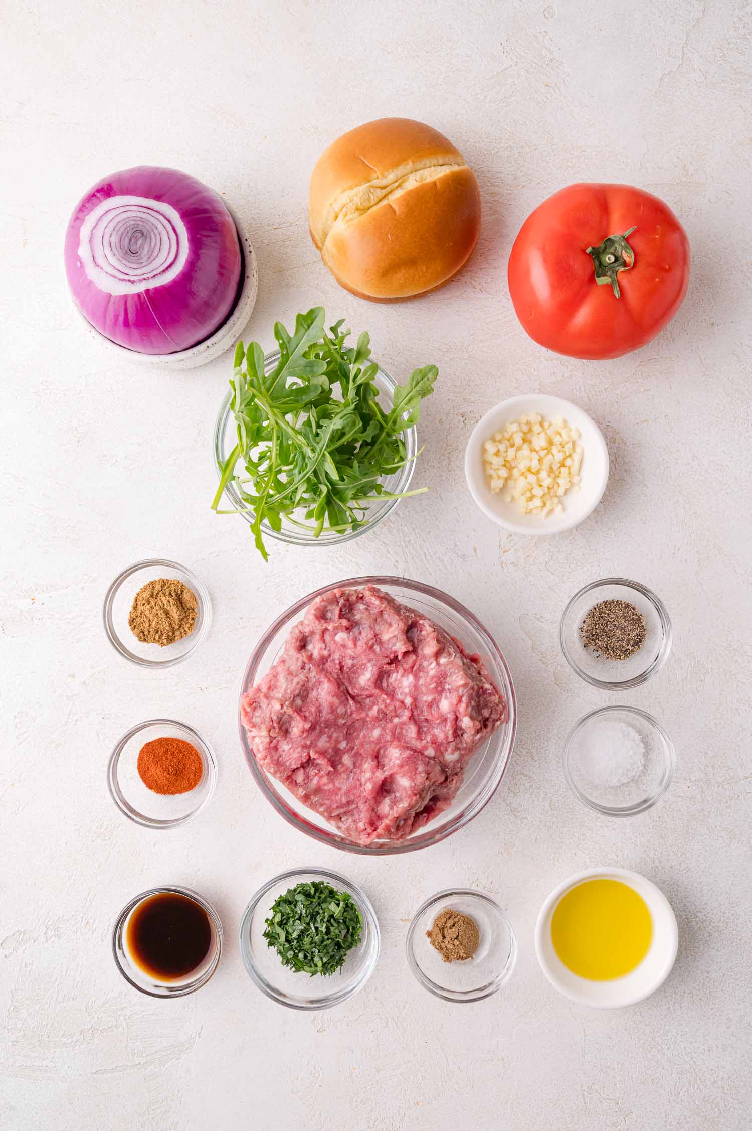 Ingredients for making burgers, including ground lamb, a bun, tomato, red onion, arugula, minced garlic, spices, herbs, Worcestershire sauce, olive oil, salt, and pepper, arranged on a white surface.