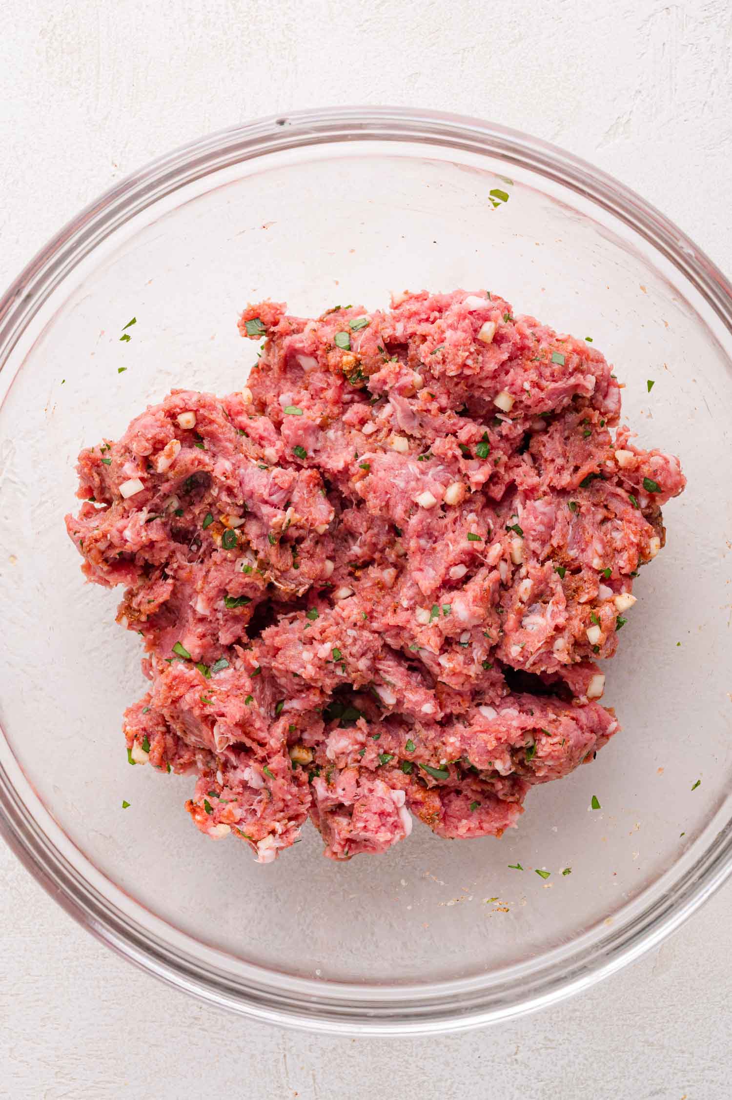 A glass bowl filled with seasoned raw ground lamb mixed with herbs and spices on a light-colored surface.
