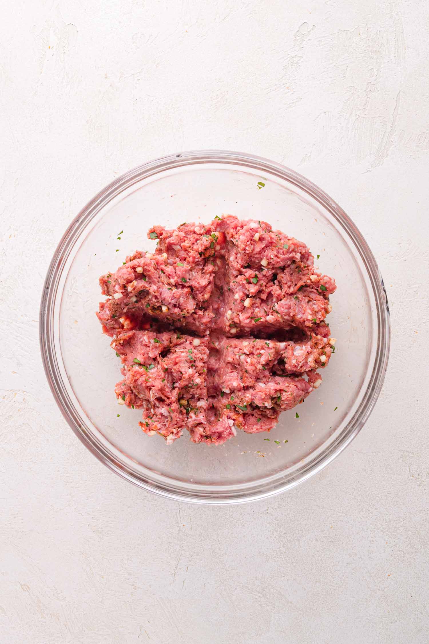 A glass bowl contains seasoned raw ground lamb divided into four equal sections on a light-colored surface.