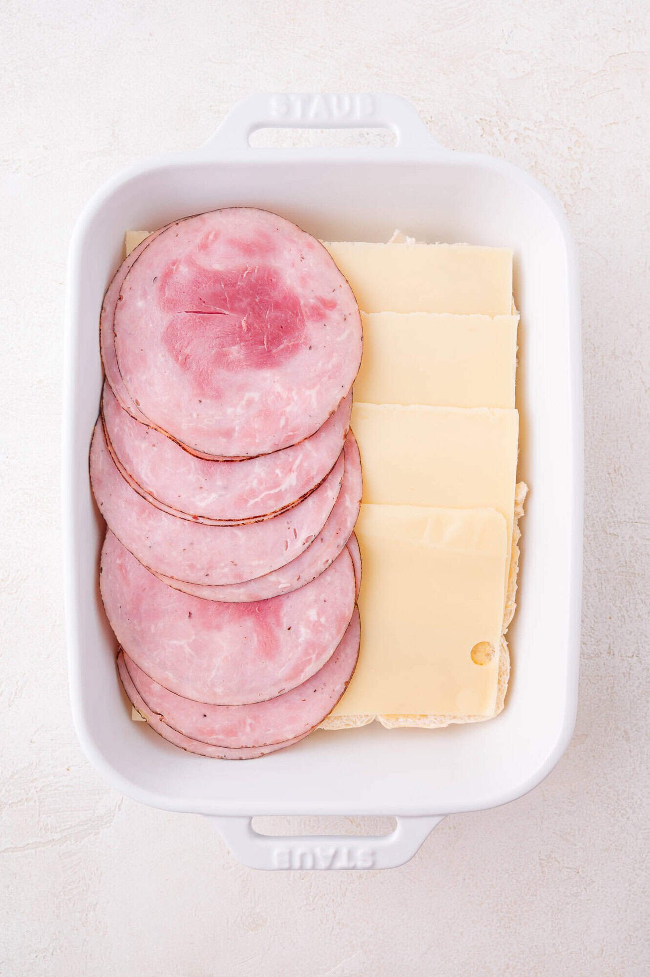 A white baking dish containing overlapping slices of ham and Swiss cheese layered over rolls.