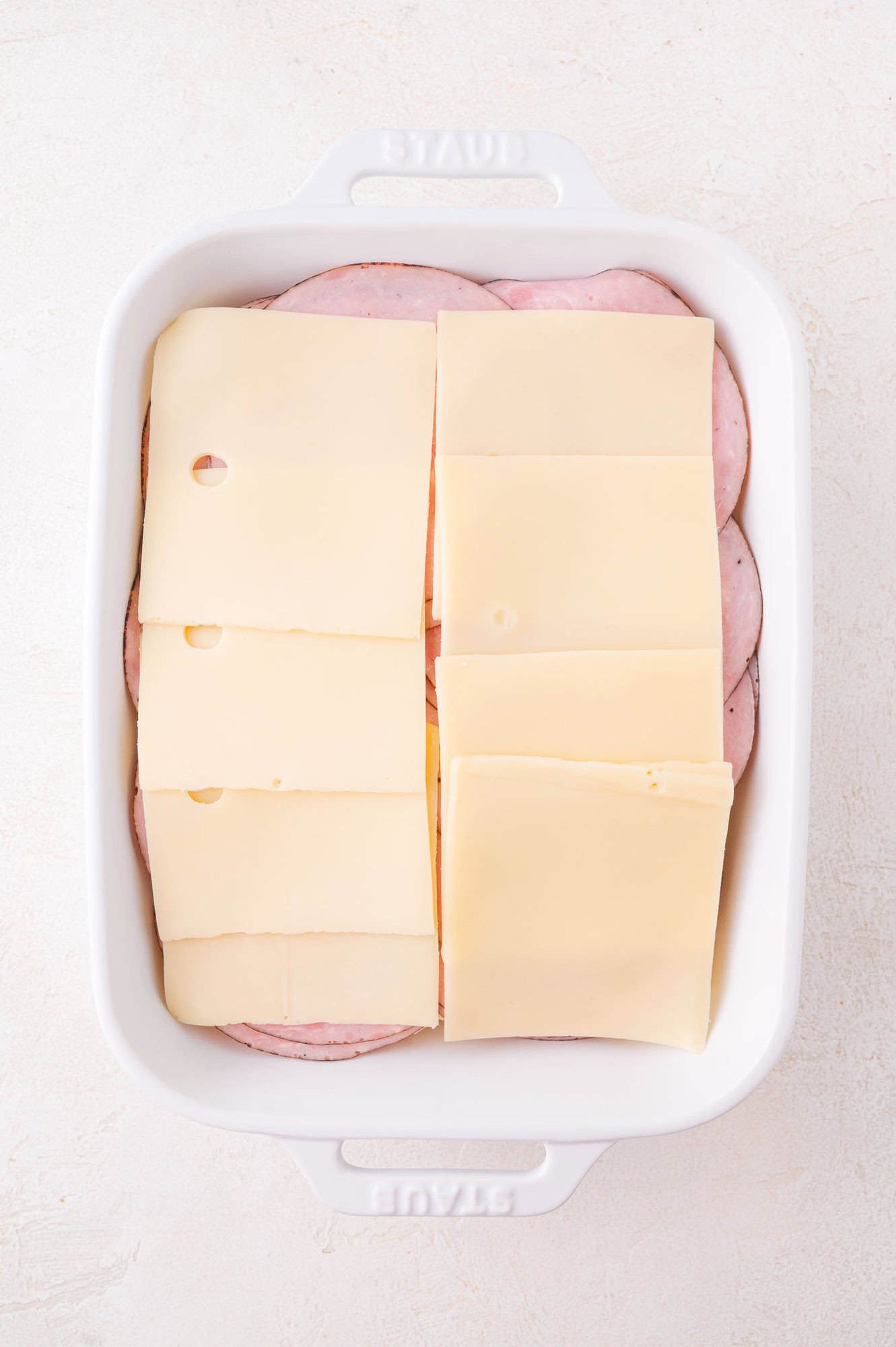 A white baking dish filled with overlapping slices of ham on the bottom and layers of Swiss cheese on top.