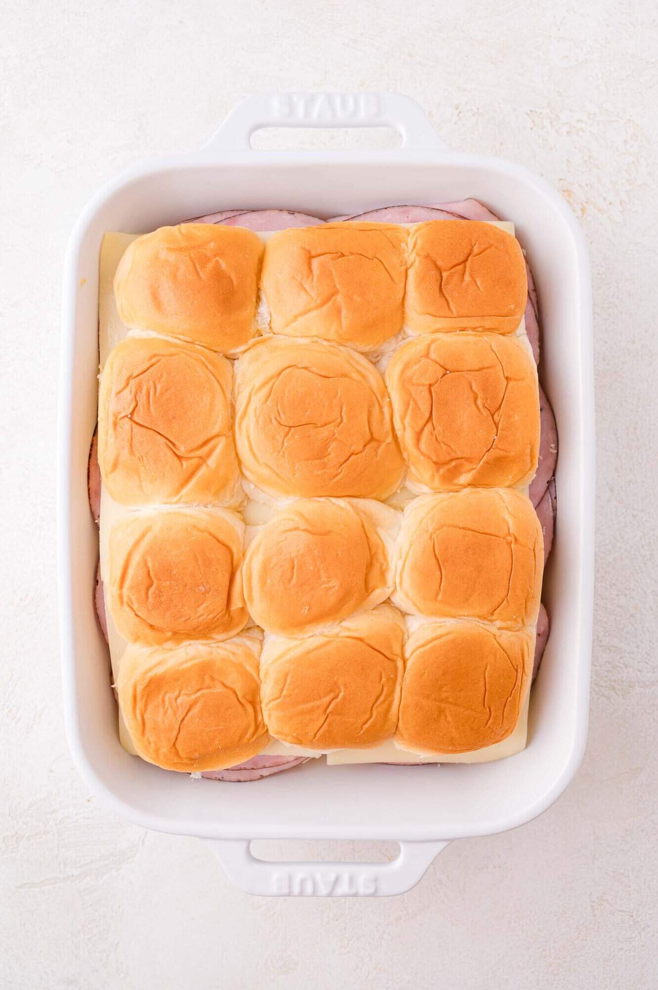A white baking dish filled with assembled slider sandwiches, featuring a top layer of golden brown buns.