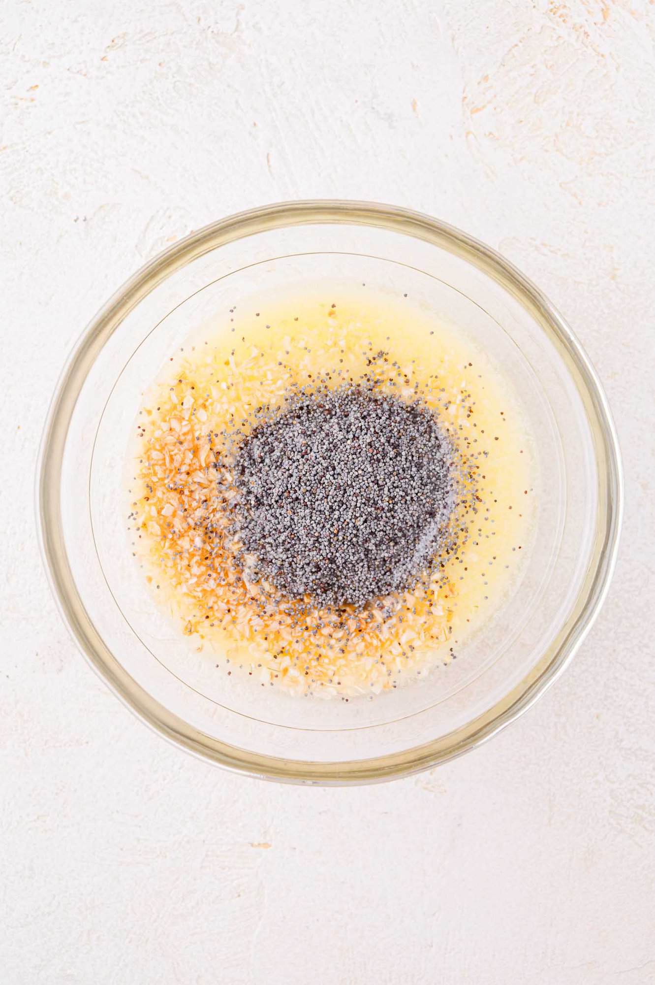 A clear glass bowl containing a mixture of poppy seeds, minced onions, and liquid ingredients on a light surface.