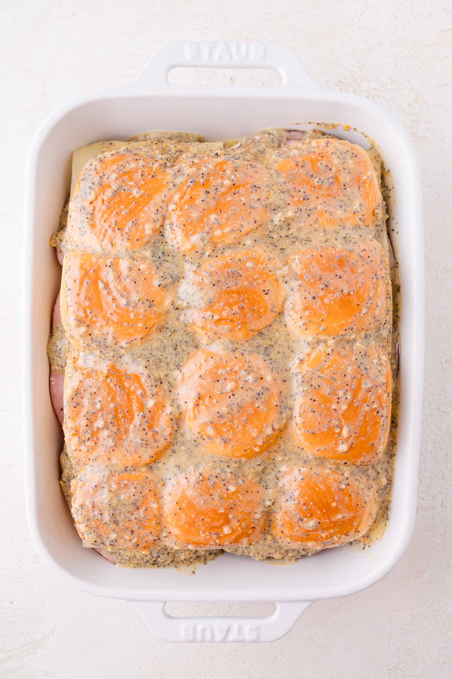 A white baking dish filled with slider sandwiches topped with a poppy seed and seasoned sauce, ready to be baked.
