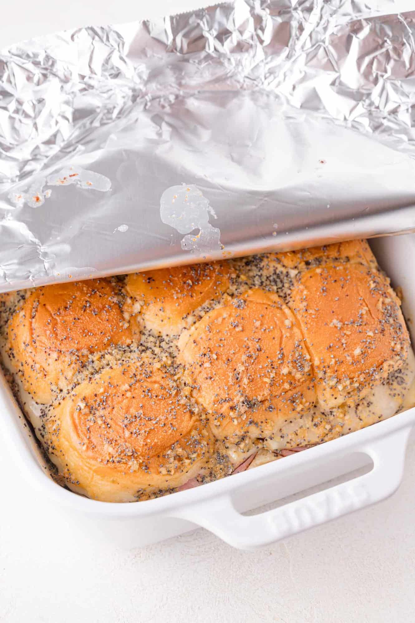 A white baking dish with poppy seed-topped buns partially covered by a sheet of aluminum foil.