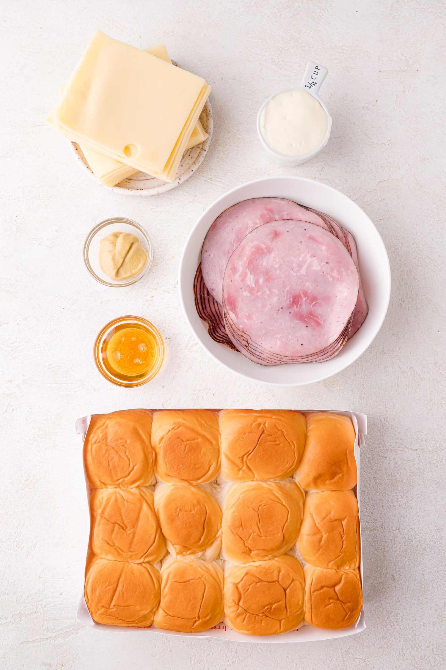 A tray of sandwich rolls next to plates of sliced ham, Swiss cheese, a bowl of mayonnaise, and small bowls of mustard and honey on a white surface.
