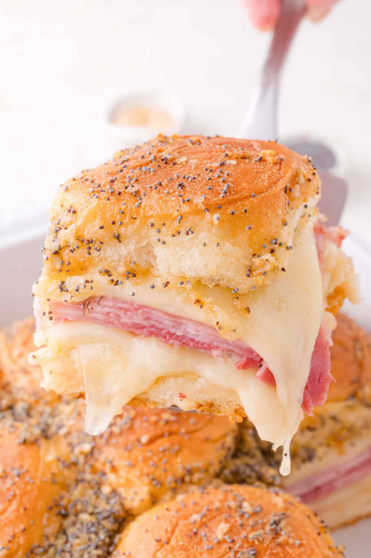 A close-up of a ham and cheese slider with poppy seeds on top, being lifted from a tray with melted cheese stretching.