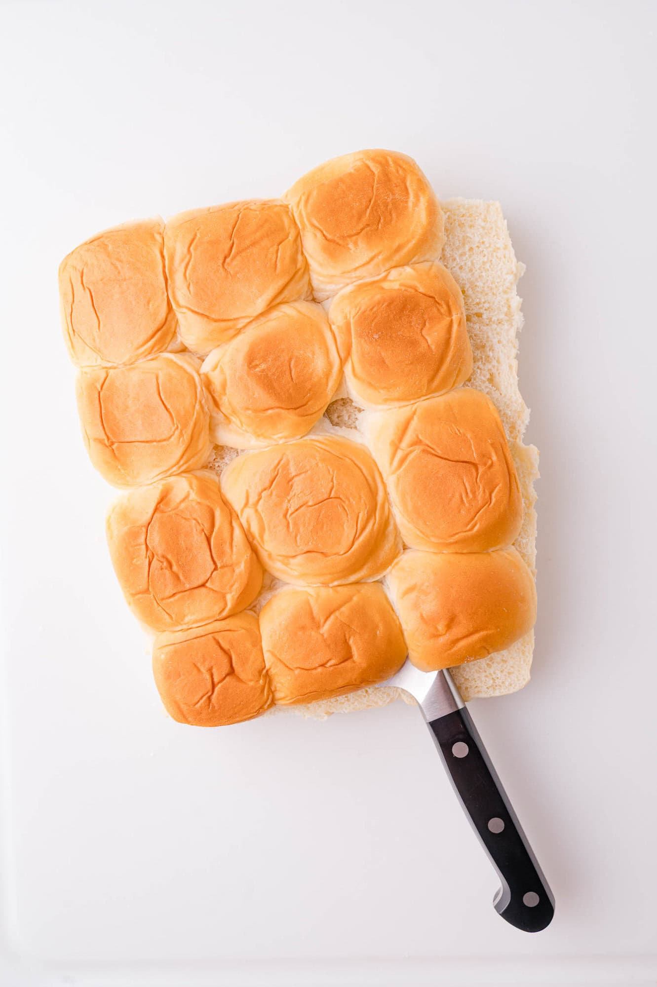 A serrated knife partially slides under a slab of twelve connected dinner rolls on a white surface.