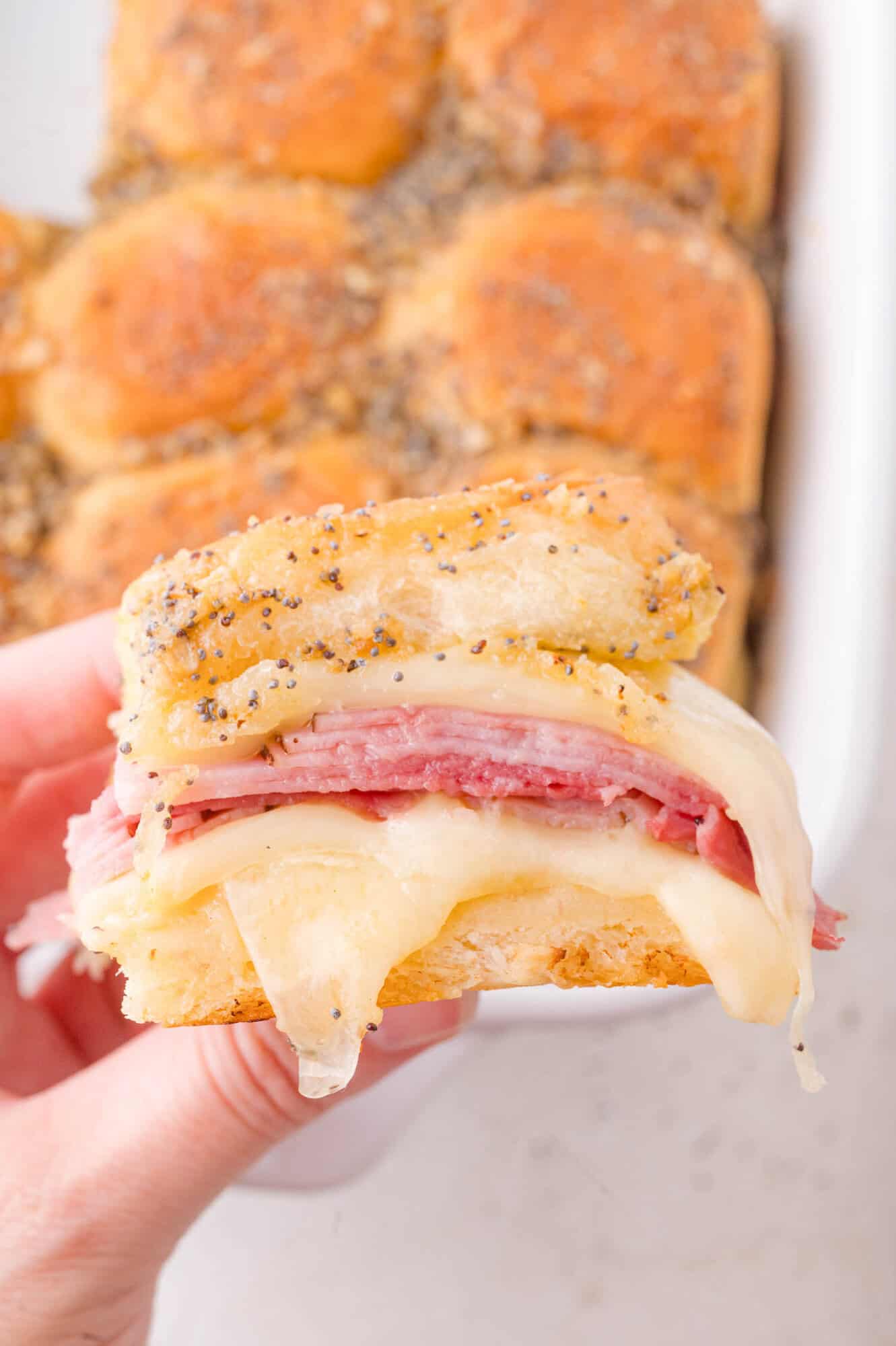 A hand holds a sandwich slider with layers of ham and melted Swiss cheese on a poppy-seed topped bun; more sliders are in the background.