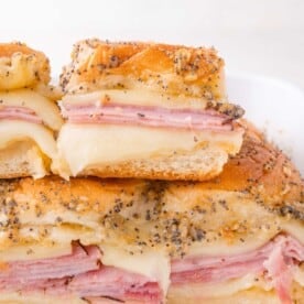 Close-up of ham and cheese sliders on soft buns, topped with poppy seeds and a buttery glaze, stacked on a white plate.