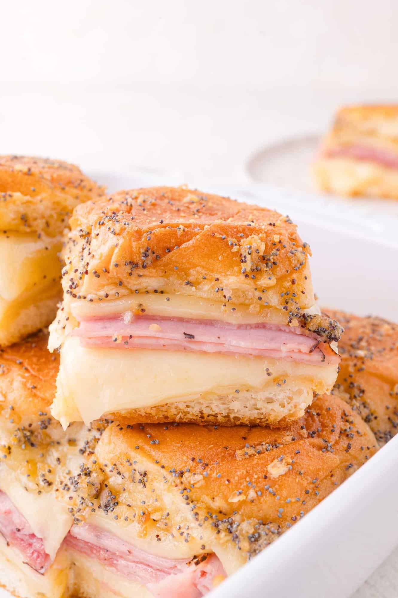 A close-up of stacked baked ham and cheese sliders with poppy seeds and seasoning on top, served in a white dish.