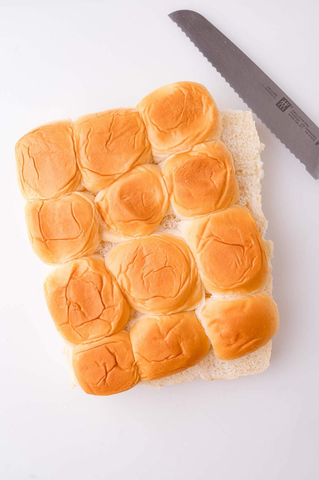 A pack of twelve sliced slider buns next to a serrated bread knife on a white surface.