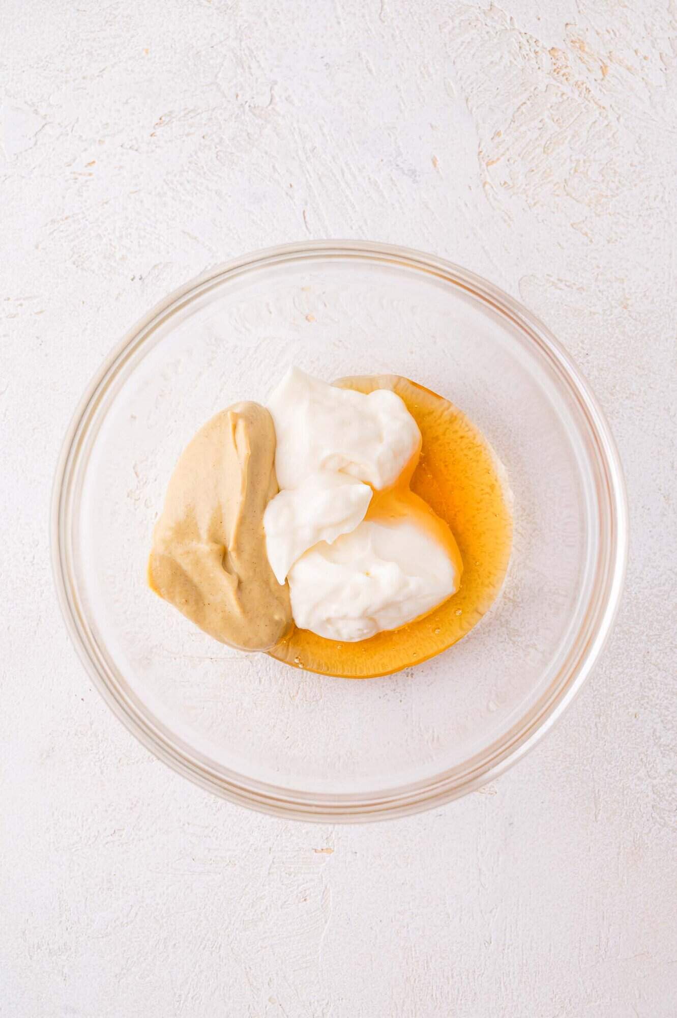 A glass bowl containing mayonnaise, mustard, and honey on a light textured surface.