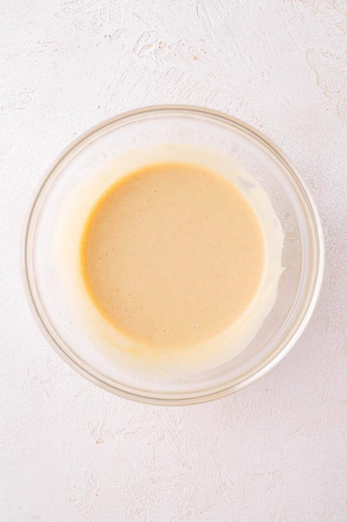 A clear glass bowl containing a light beige batter or sauce sits on a textured white surface.