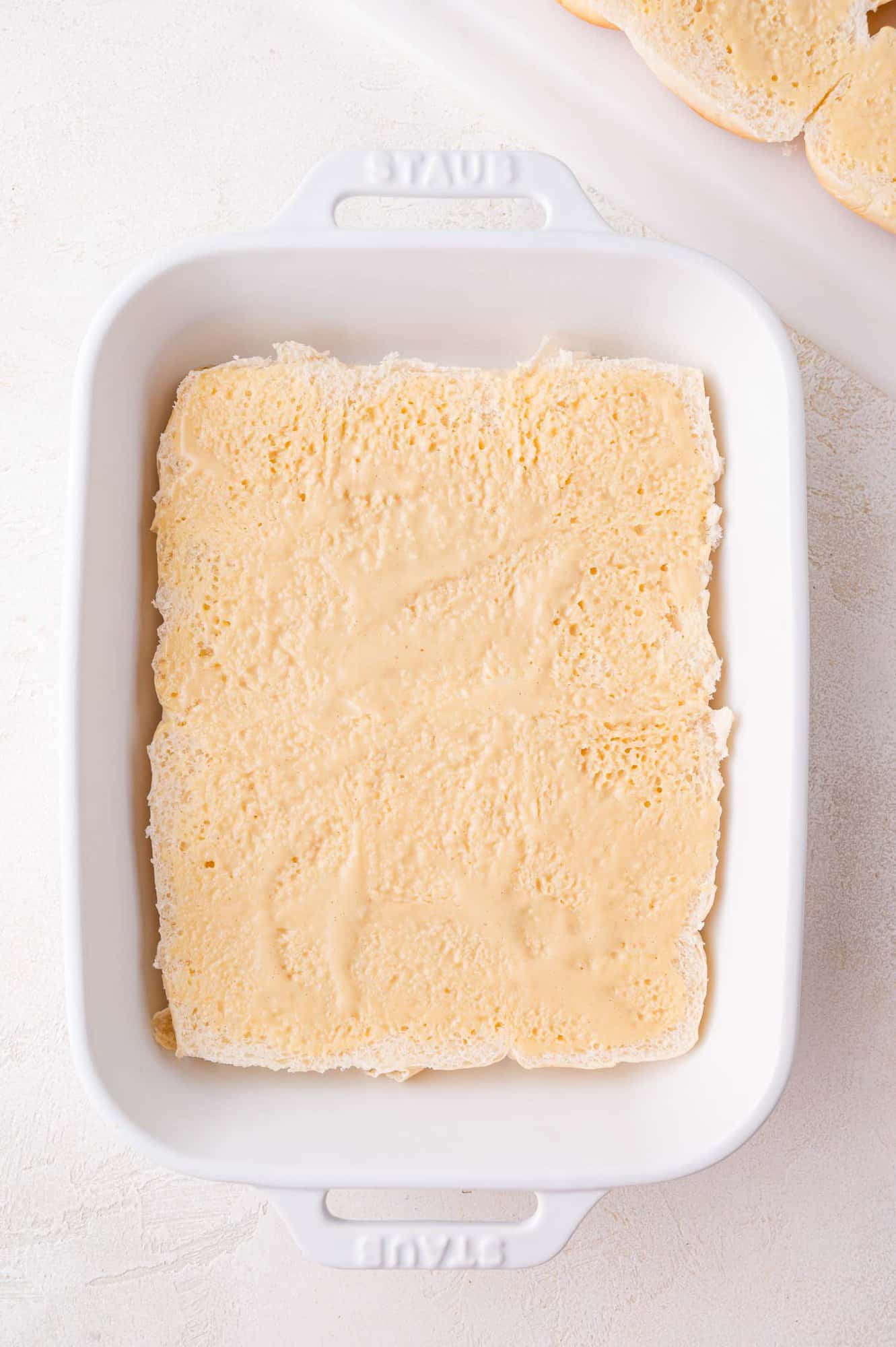 A white baking dish containing a single layer of pale, spongy rolls, topped with mayo and mustard mixture.