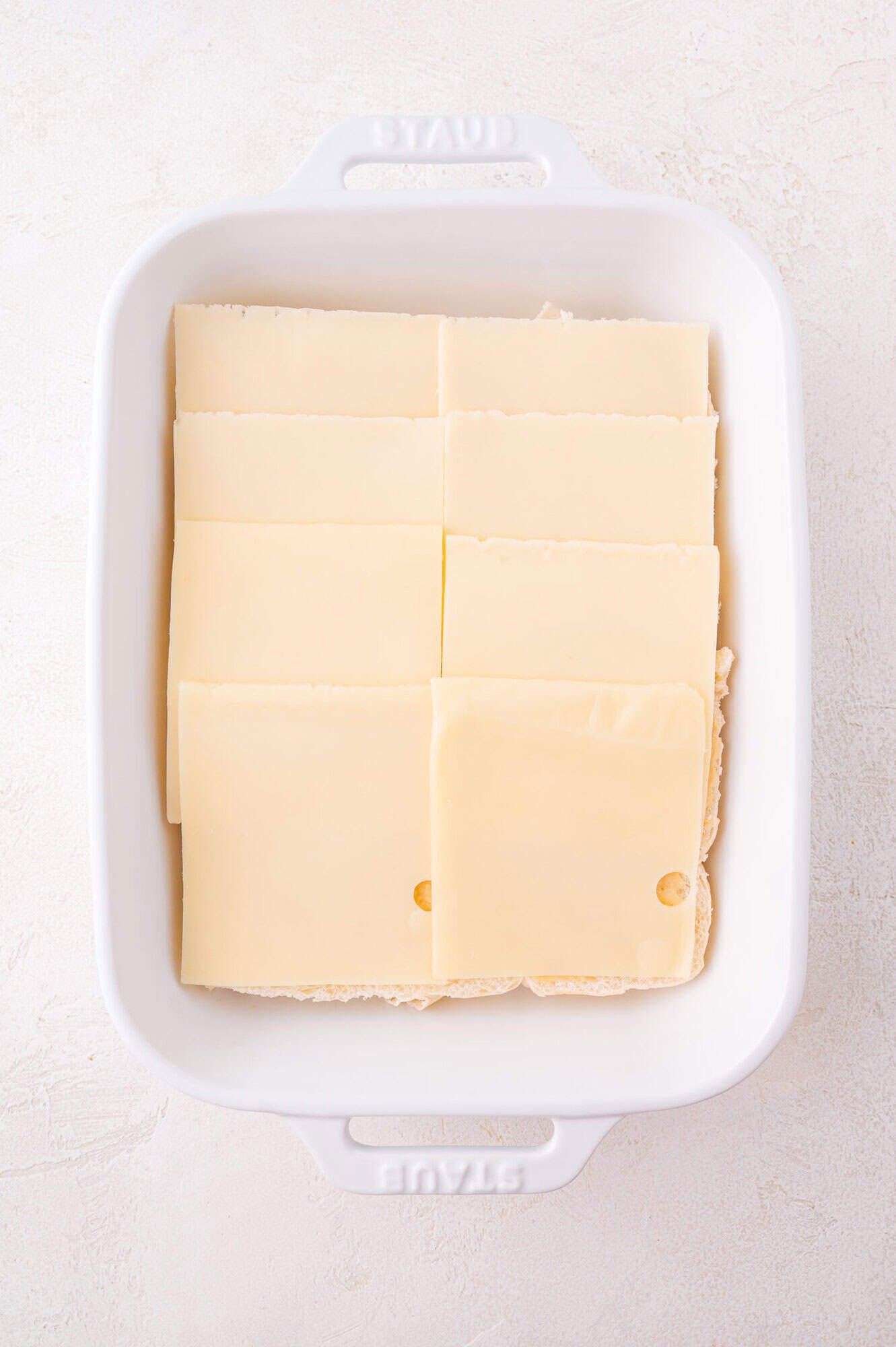 A white baking dish with neatly arranged slices of Swiss cheese covering the rolls on the bottom.
