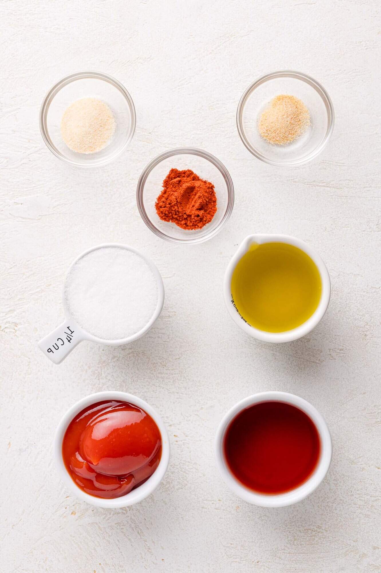 Seven small bowls on a white surface contain garlic powder, onion powder, paprika, sugar, olive oil, ketchup, and vinegar, arranged in a neat pattern.