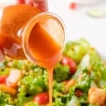 A hand pours orange Catalina salad dressing from a jar onto a fresh green salad with lettuce, tomatoes, and croutons.