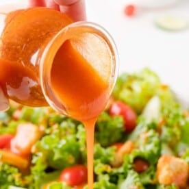 A hand pours orange Catalina salad dressing from a jar onto a fresh green salad with lettuce, tomatoes, and croutons.