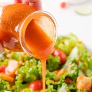 A hand pours orange Catalina salad dressing from a jar onto a fresh green salad with lettuce, tomatoes, and croutons.