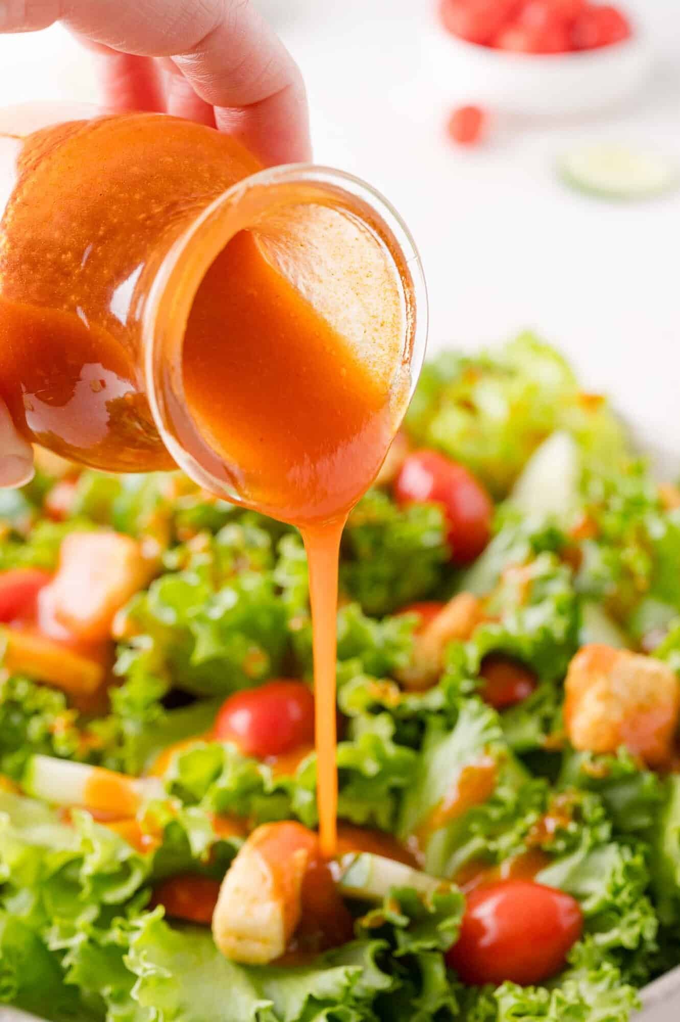 Hand pouring orange colored Catalina dressing from a small jar onto a fresh green salad with lettuce, cherry tomatoes, and croutons.