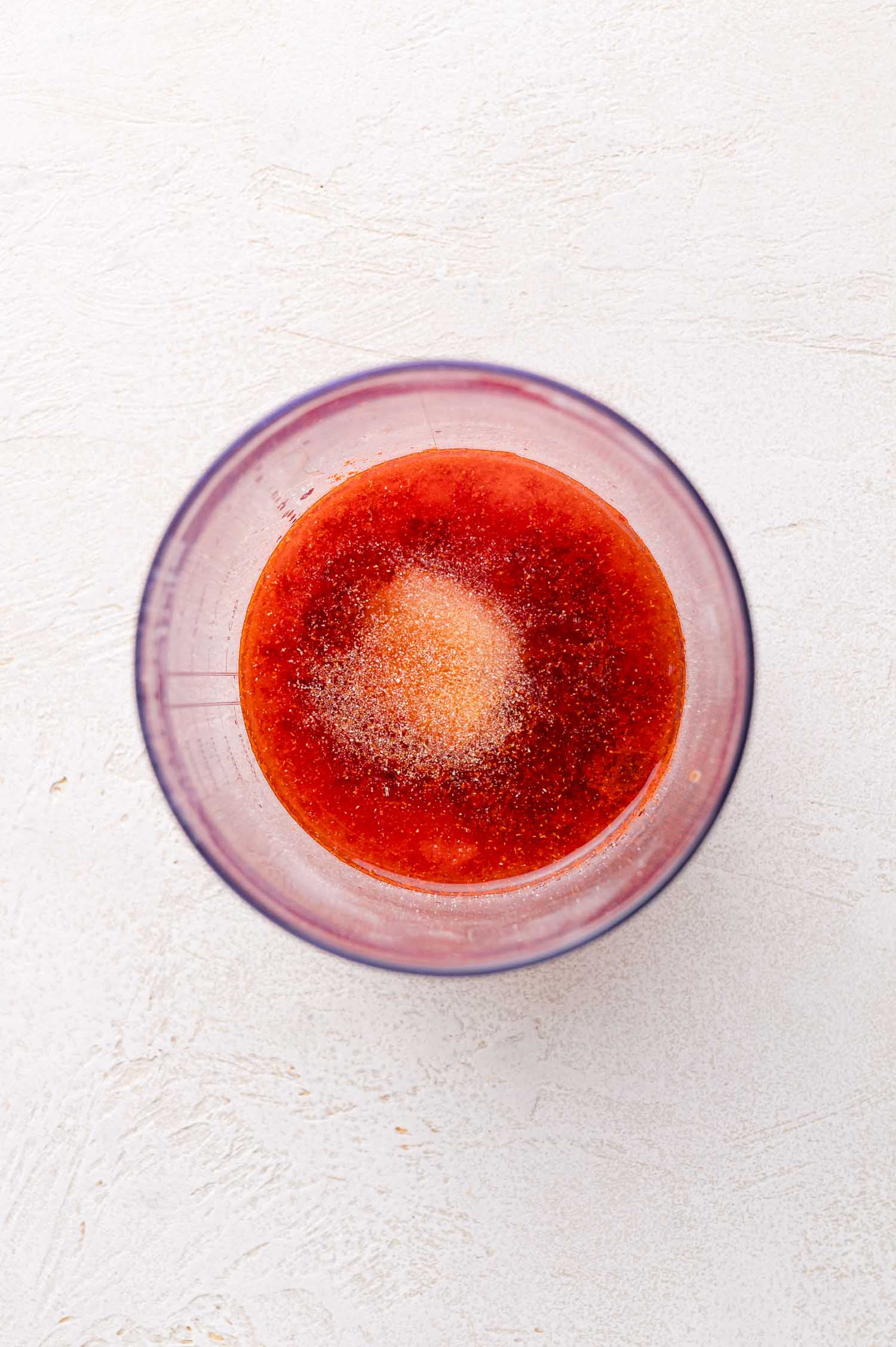 A clear bowl containing a red liquid mixture with a mound of granulated seasoning in the center, viewed from above on a light textured surface.