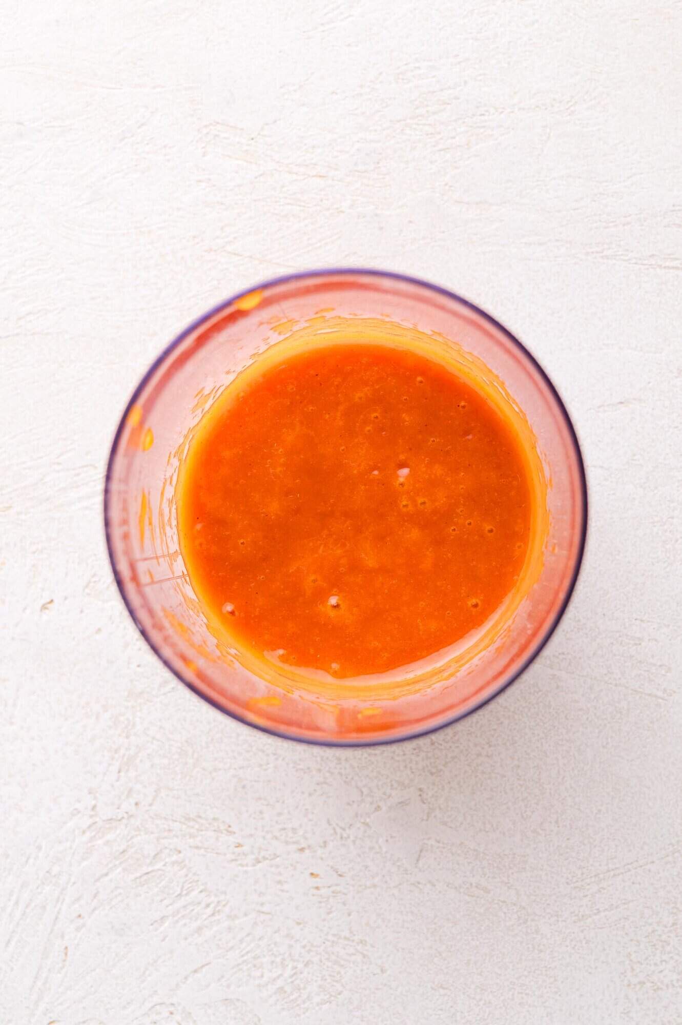 A clear glass bowl filled with smooth, bright orange salad dressing on a white textured surface, viewed from above.