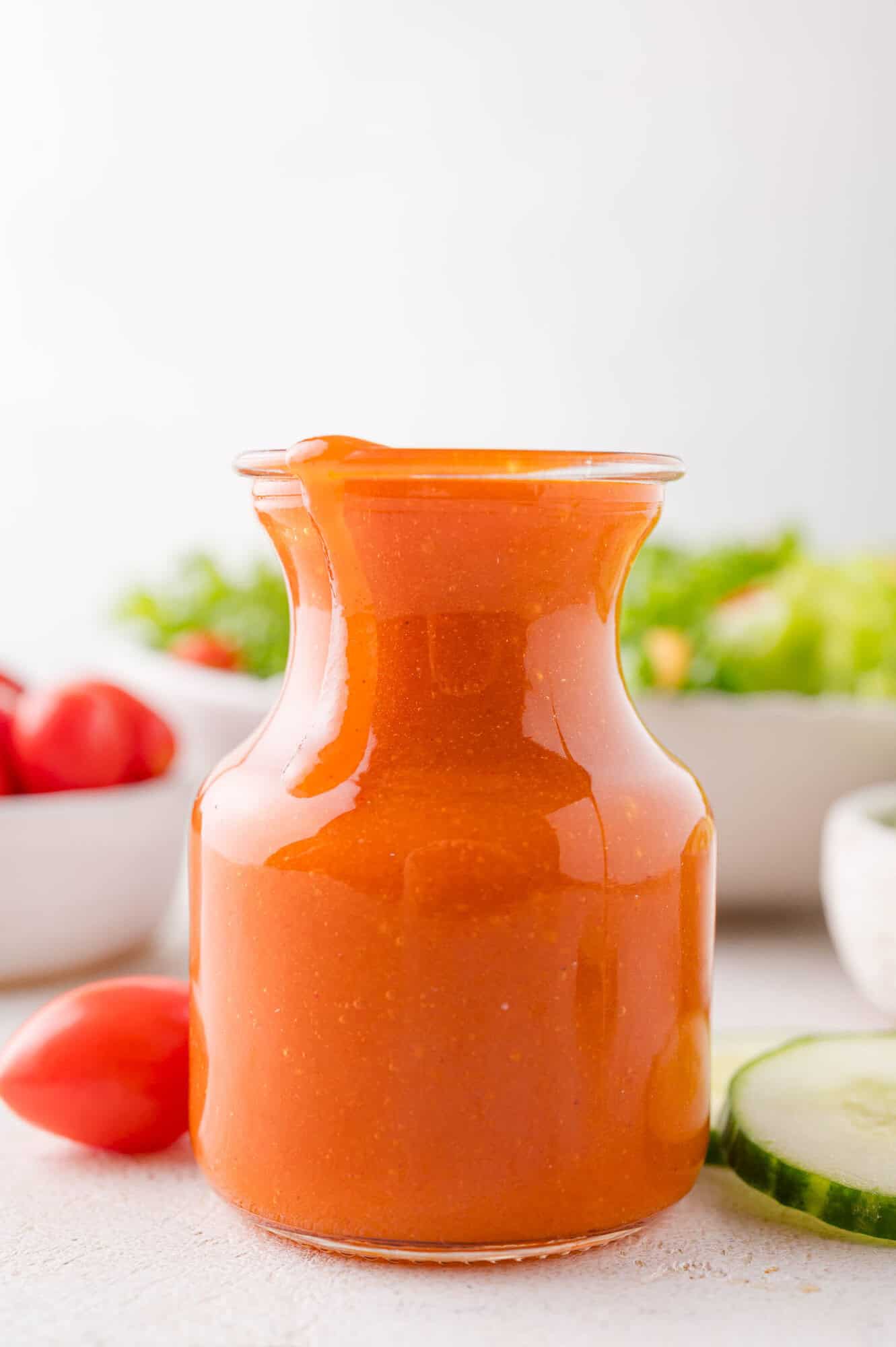 A glass jar filled with orange colored salad dressing sits on a white surface, with vegetables like tomatoes and cucumber in the background.