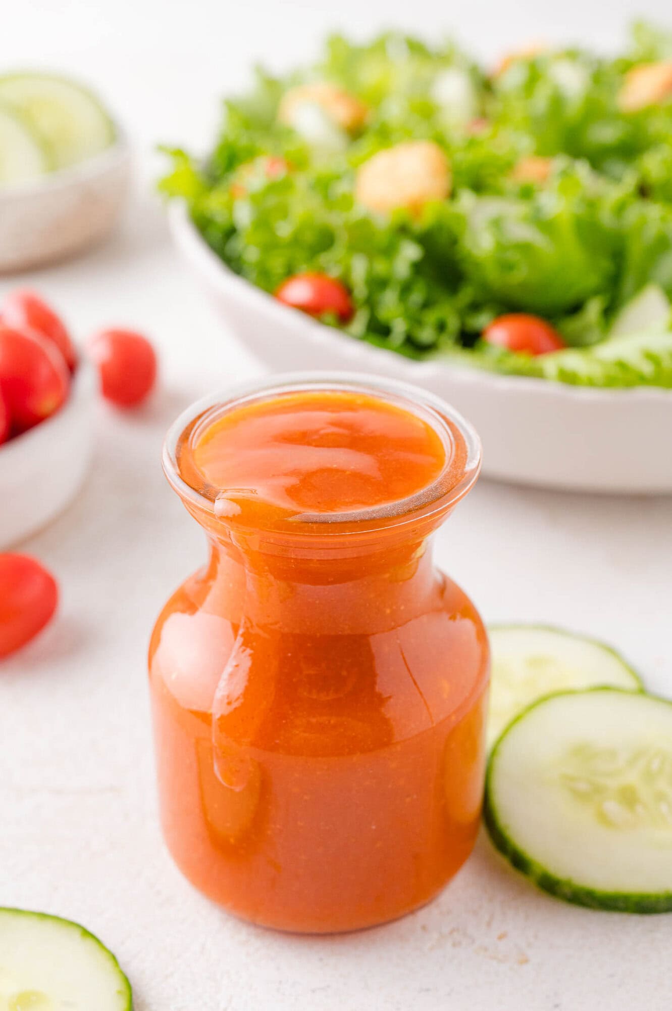 A glass jar filled with Calalina salad dressing sits on a white surface, with cucumber slices and a bowl of green salad in the background.