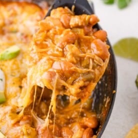 A serving of cheesy enchilada skillet being lifted from a casserole dish with a spoon, showing melted cheese stretching.