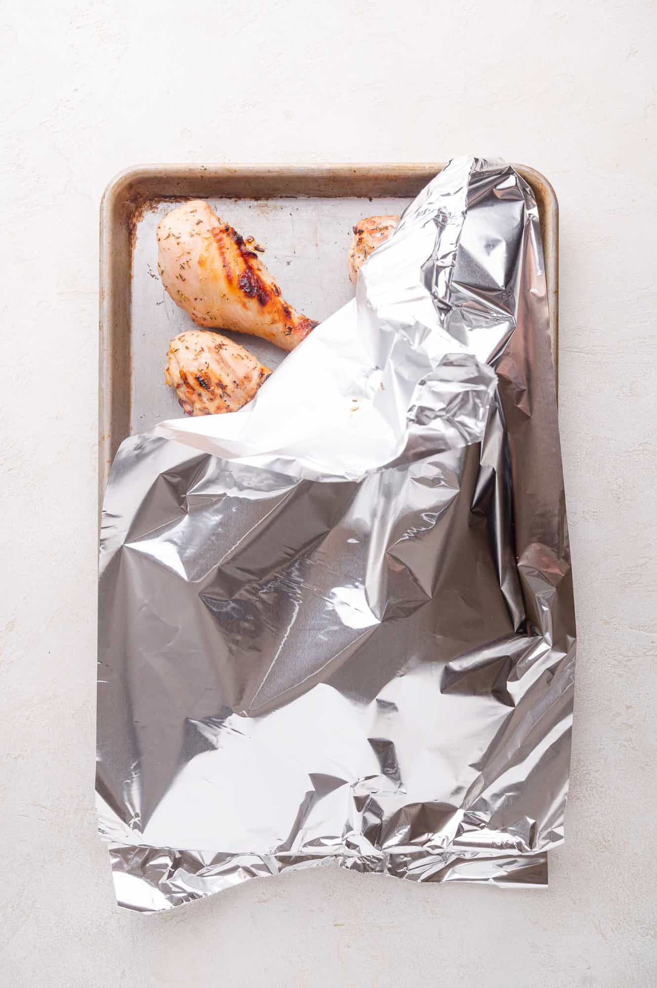 A baking sheet with cooked chicken drumsticks partially covered by a sheet of aluminum foil on a light surface.
