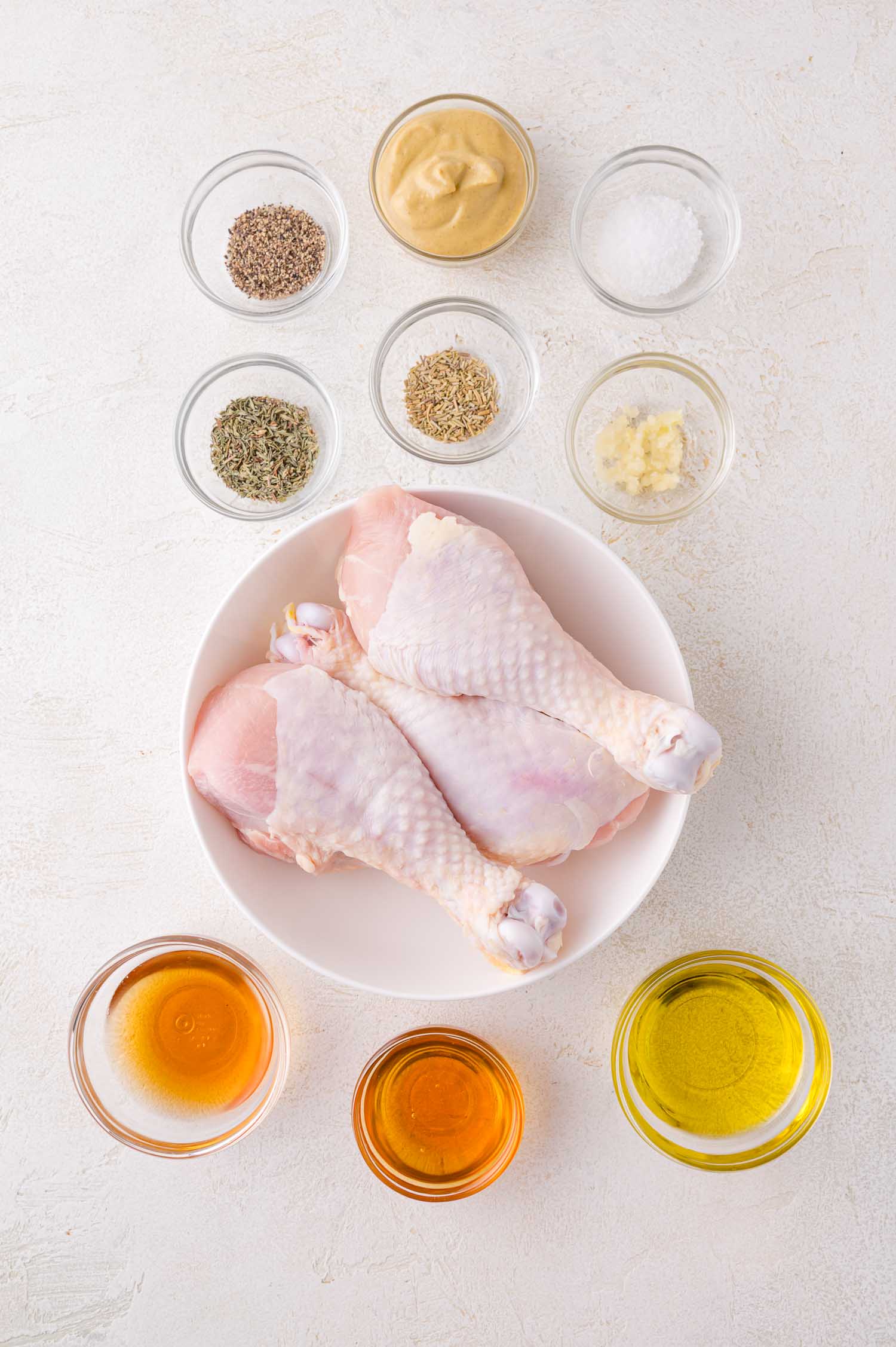 A bowl of raw chicken drumsticks surrounded by small bowls of various spices and seasonings, arranged on a white surface.