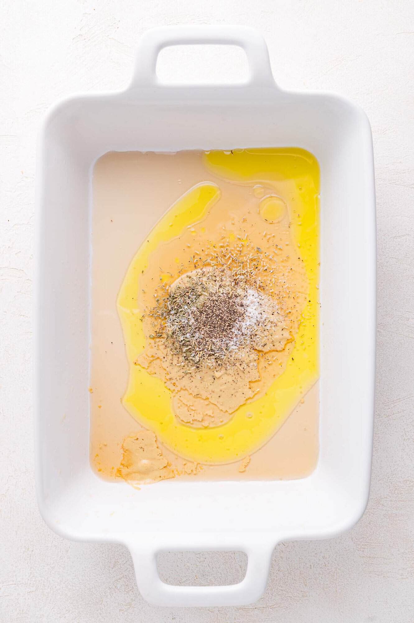 A white baking dish containing a not yet mixed marinade on a white surface.