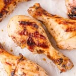 Close-up of several grilled chicken drumsticks with a golden-brown, slightly charred exterior on a sheet of parchment paper.